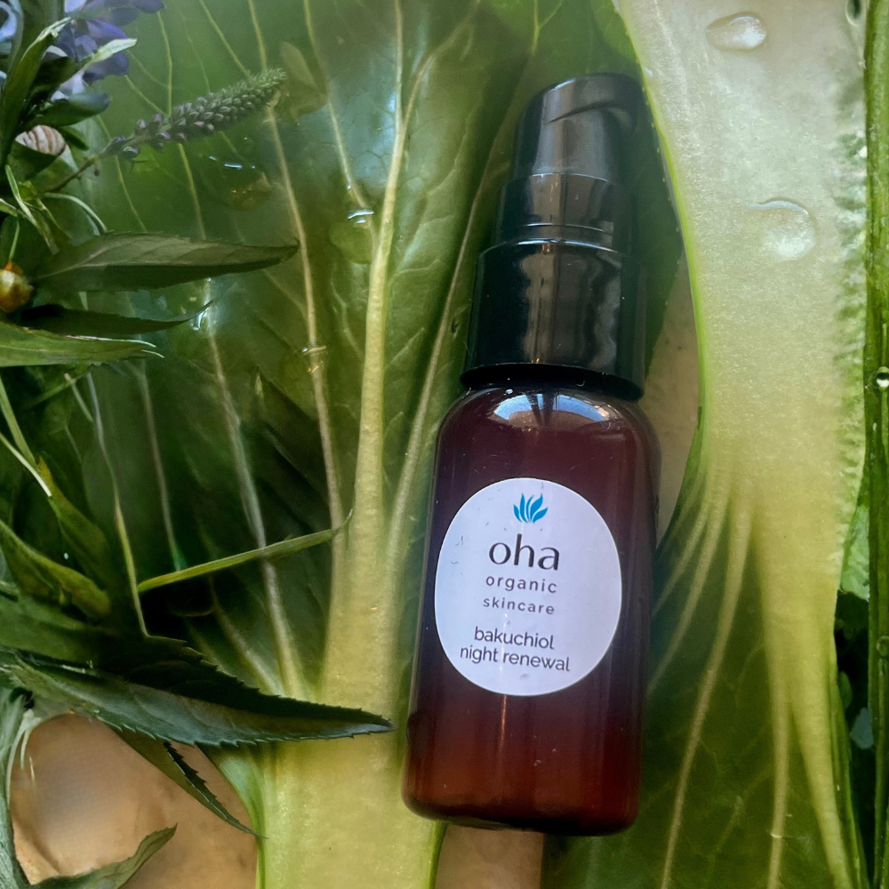 A bottle of oha organic skincare bakuchiol night renewal serum, placed on large green leaves with water droplets