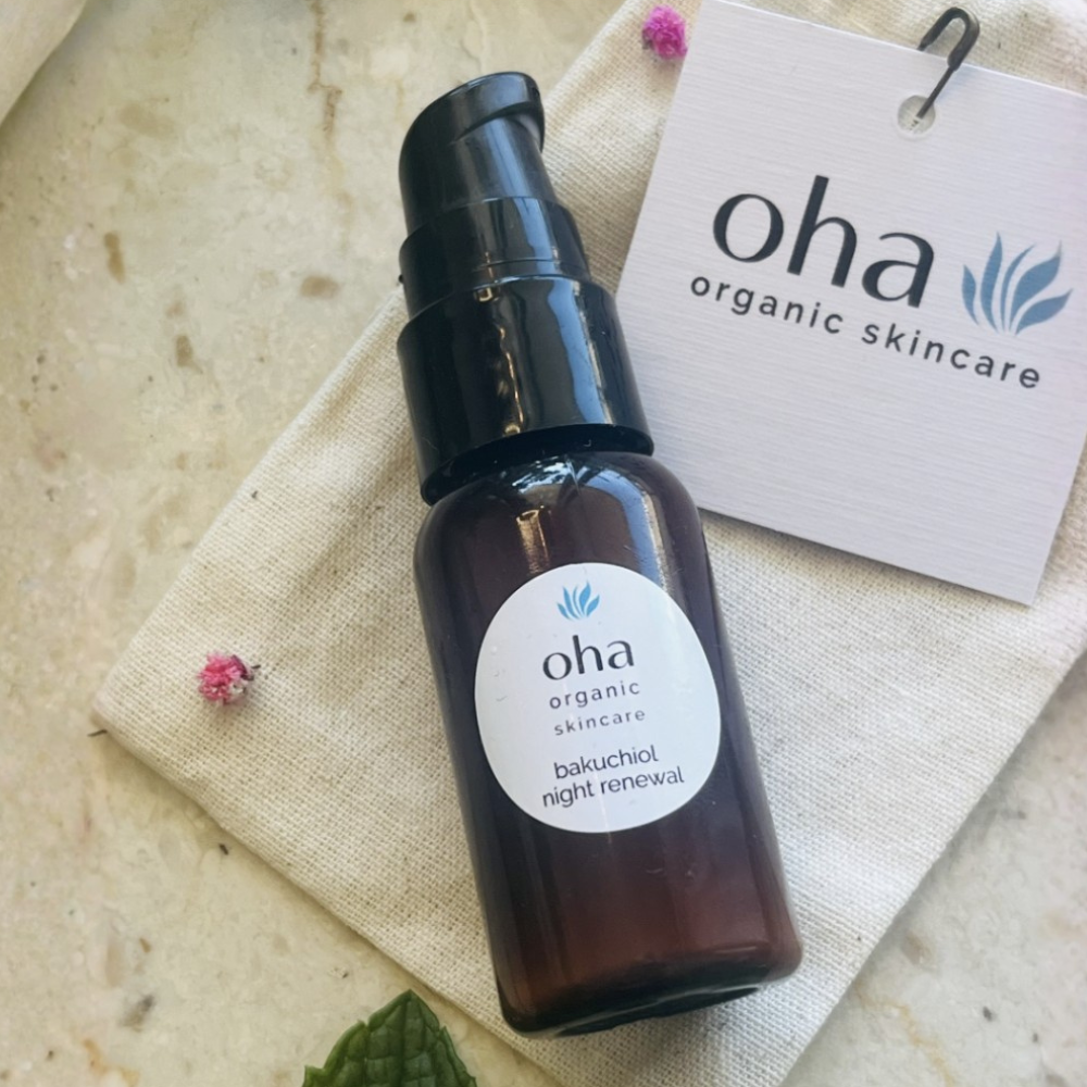 A dark amber glass pump bottle labeled 'oha organic skincare bakuchiol night renewal' resting on a beige fabric bag with a branded white hangtag