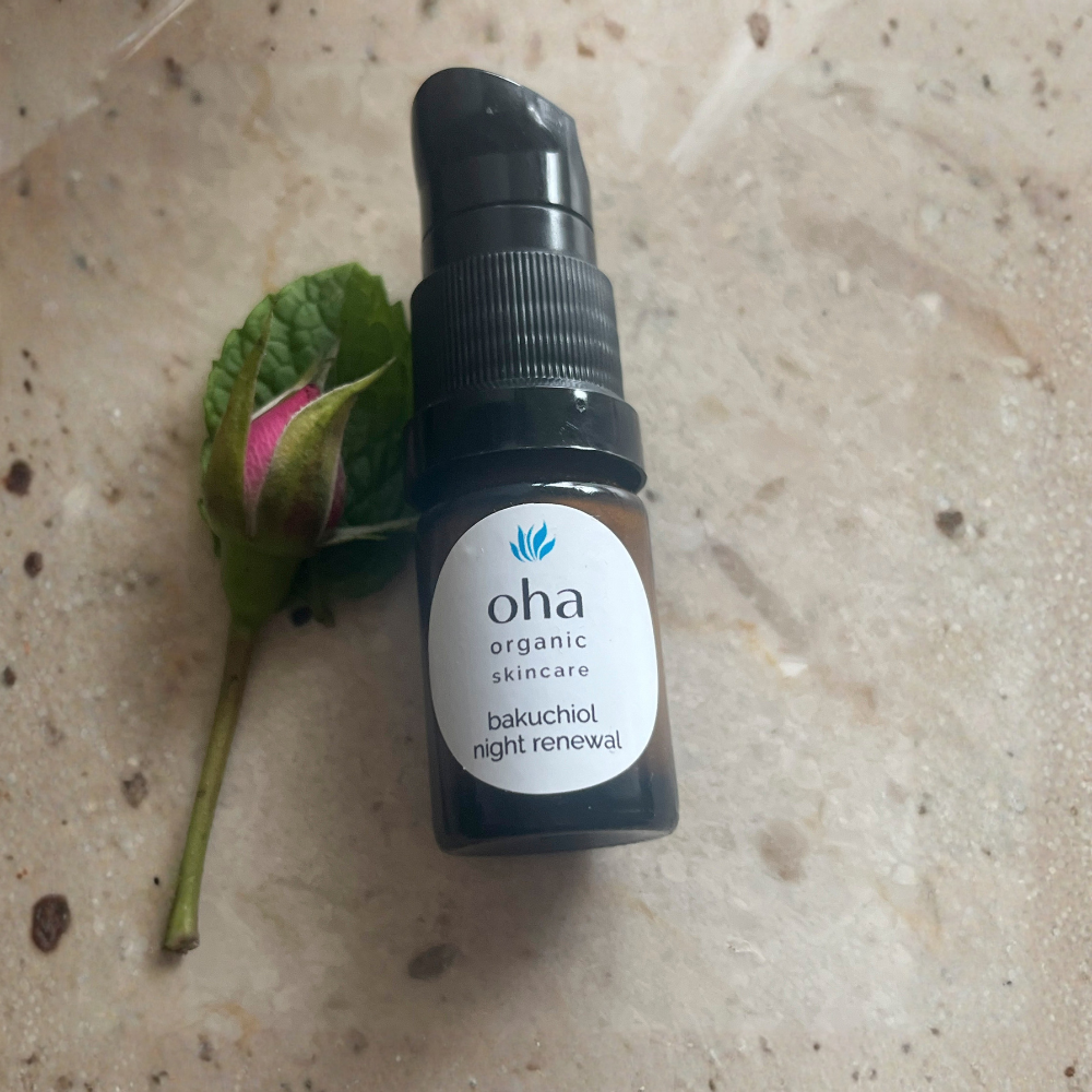A small sample size of bakuchiol night renewal in amber glass pump bottle labeled 'oha organic skincare  sits on a light beige stone surface next to a green leaf and pink bud.